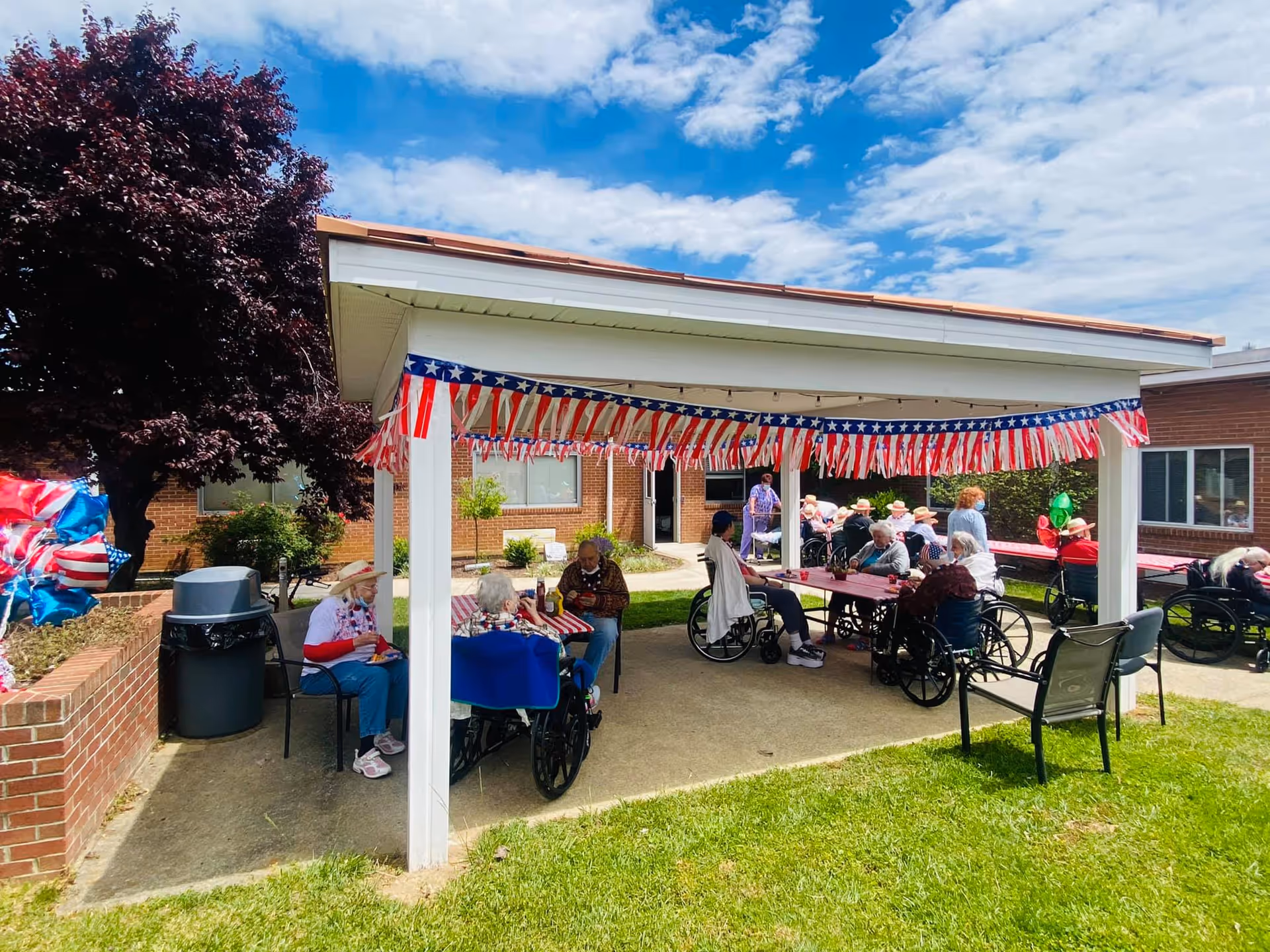 Outdoor covered patio area at a nursing home with several elderly residents sitting at tables and in wheelchairs, decorated with red, white, and blue American flag-themed garlands. The sky is partly cloudy and the building is made of brick.