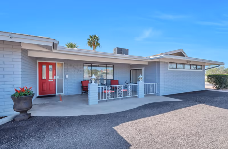 Exterior view of a single-story building with light gray brick walls and a red front door. There is a small covered patio area with red cushioned chairs and a table, bordered by a white metal railing. A large planter with red flowers is near the entrance. The sky is clear and blue with a palm tree visible in the background.