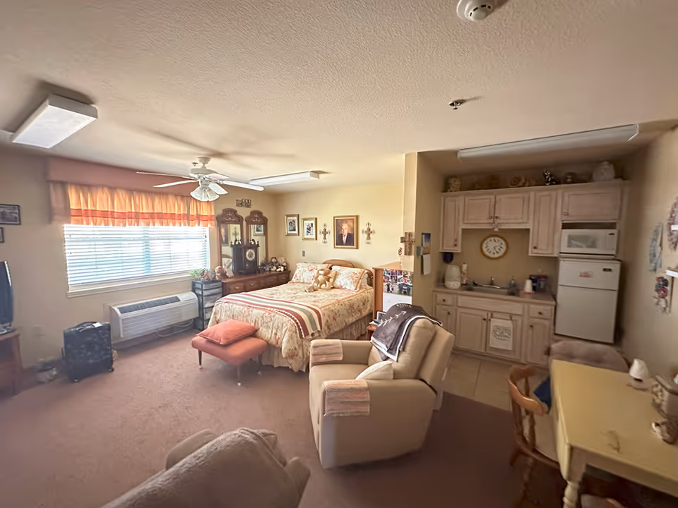 Studio-style assisted living room with a bed, recliner chairs, dresser under a window, and a small kitchenette area.