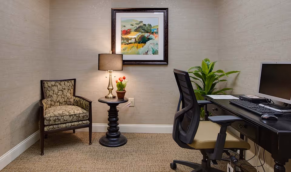 A small office corner with a black desk holding a computer monitor, keyboard, mouse, and some papers. There is a black office chair with a beige seat in front of the desk. To the left, a round black side table with a lamp and a small potted plant with red flowers sits next to a patterned armchair. A framed colorful landscape painting hangs on the beige wall above the side table. A green leafy plant is placed behind the desk near the wall.