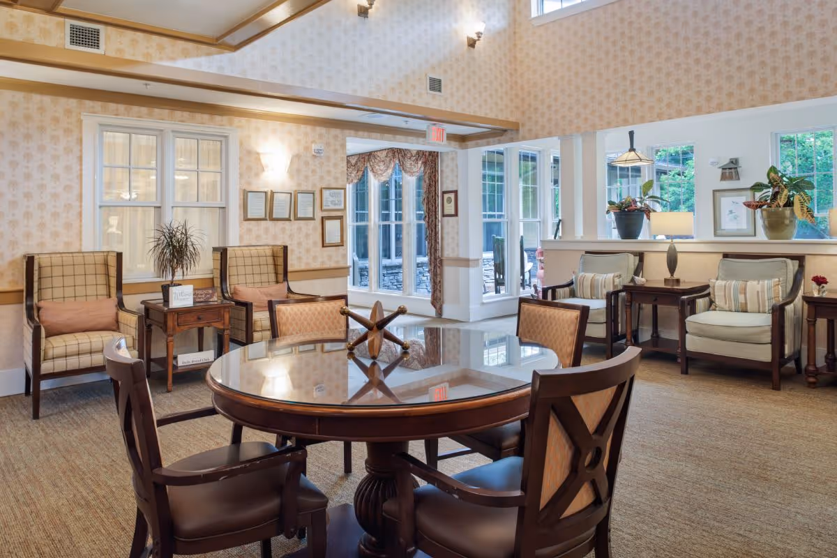 Cozy senior living common room with a round wooden table, chairs, armchairs, and large windows letting in natural light.