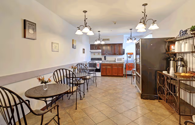 A dining area with several round metal tables and chairs along the left wall, each table decorated with a small vase of flowers. The room has tiled floors and white walls adorned with framed coffee-themed artwork. At the far end, there is a kitchenette with wooden cabinets, a microwave, and a popcorn machine. On the right side, there is a black refrigerator and a metal rack holding coffee supplies including coffee pots, cups, and a basket of bread rolls. Multiple ceiling light fixtures illuminate the space.
