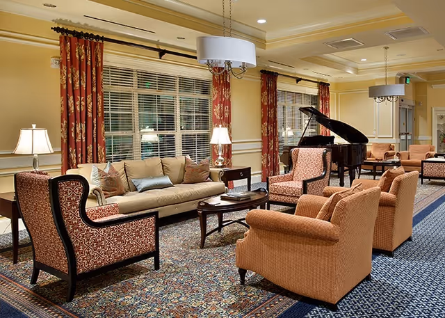A spacious and elegantly furnished living room area in a senior living facility featuring a beige sofa with multiple cushions, patterned armchairs, a wooden coffee table, a grand piano, and warm lighting from table lamps and ceiling fixtures. The room has large windows with red patterned curtains and a decorative carpet covering the floor.
