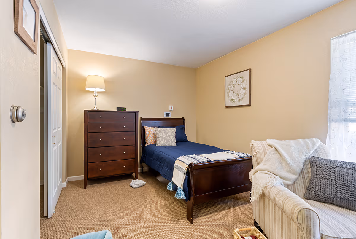 A cozy bedroom with beige walls and carpet. The room features a dark wooden bed with navy blue bedding and decorative pillows, a matching tall dresser with a lamp and digital clock on top, a striped armchair with a gray pillow and white throw blanket, and a framed floral artwork on the wall. A window with sheer white curtains allows natural light to enter the room.