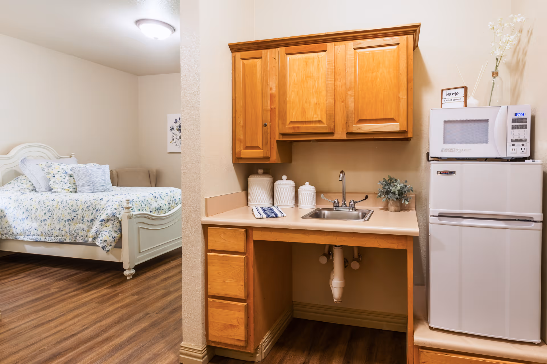 A cozy senior living studio apartment featuring a small kitchenette with wooden cabinets, a countertop with a sink, a microwave on top of a mini refrigerator, and decorative items. To the left, there is a white bed with floral bedding and multiple pillows, along with a beige armchair in the corner. The room has wood flooring and neutral-colored walls.