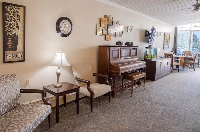 Communal lounge with chairs and a side table and lamp, an upright piano against the wall, a TV cabinet and a dining table near a window.