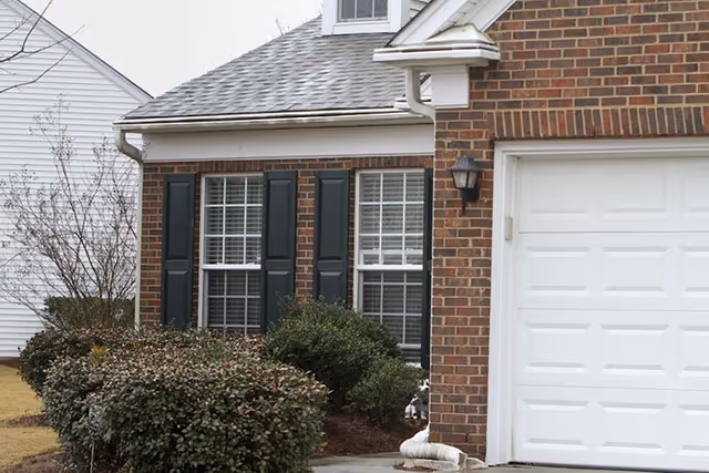 Exterior view of a brick residential building with two windows featuring black shutters and a white garage door. There are bushes and shrubs planted along the front of the building and a small tree on the left side.