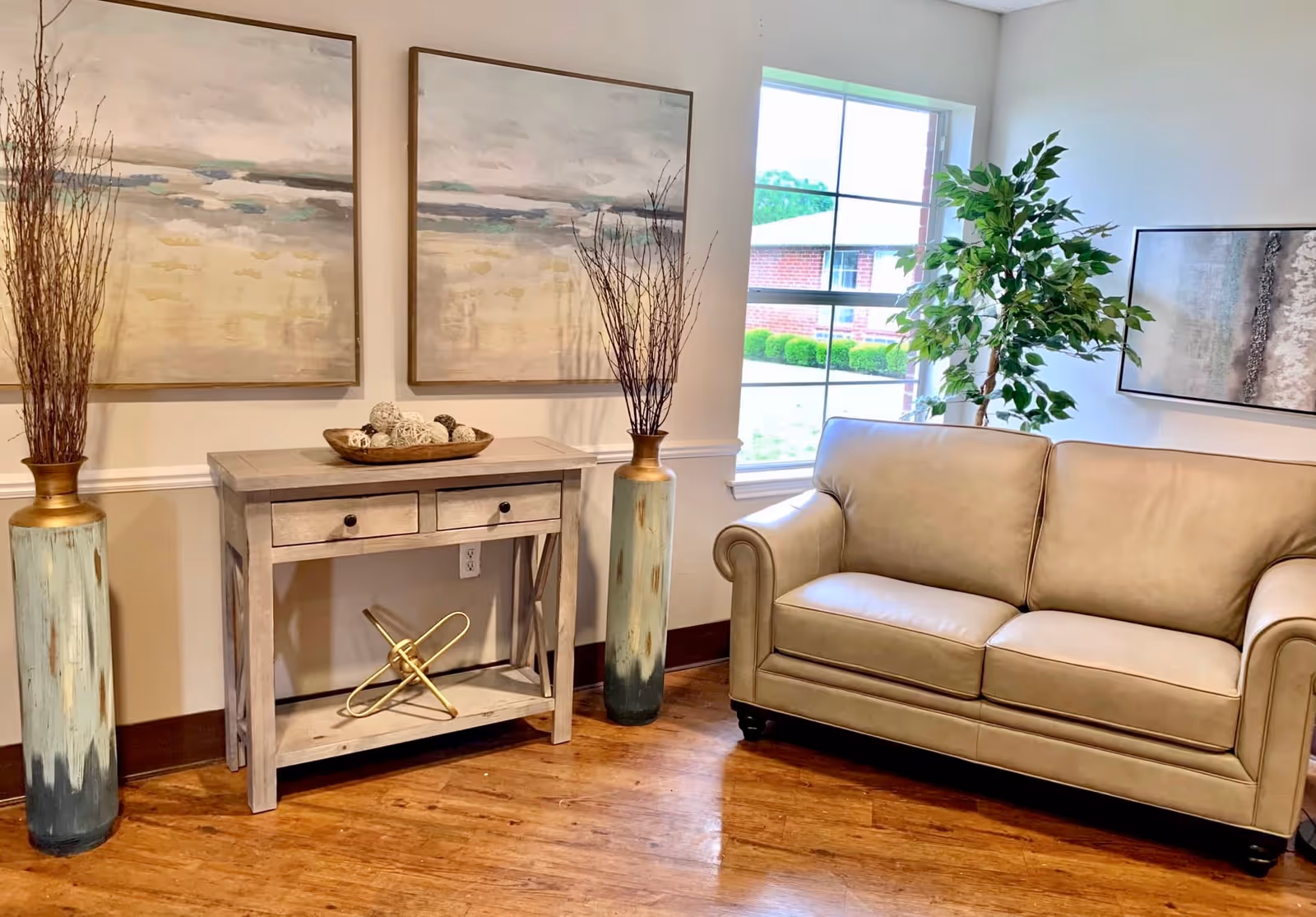 A cozy living room area with a beige leather sofa, a wooden console table with decorative items including a bowl of woven balls and a gold geometric sculpture, two tall vases with dried branches on either side of the table, two large abstract paintings on the wall, a large window showing greenery outside, and a potted plant in the corner.