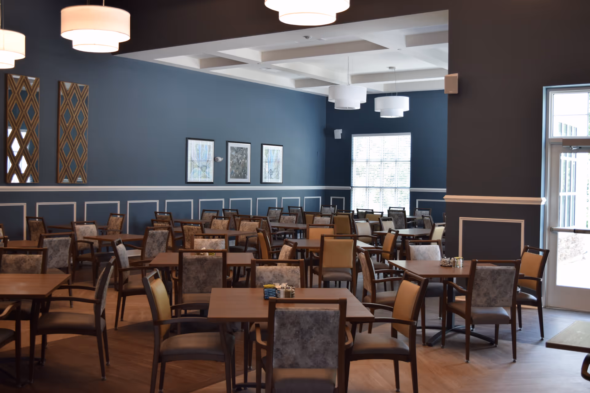 A spacious dining room with multiple wooden tables and chairs arranged neatly. The walls are painted blue with white trim and decorated with framed artwork and mirrors. Large windows allow natural light to brighten the room, and modern pendant lights hang from the ceiling.