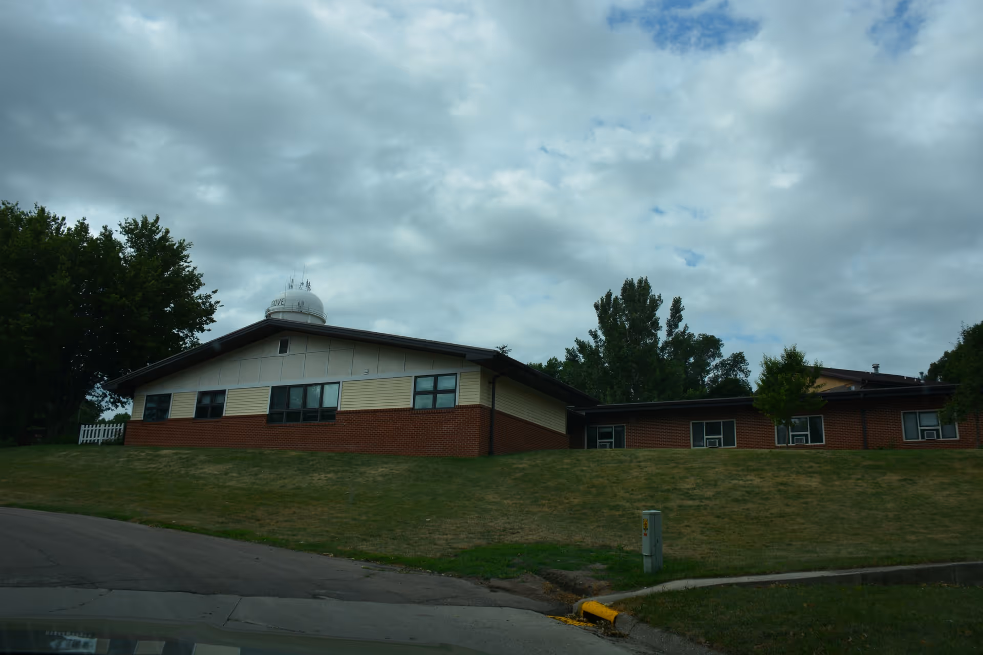 Exterior view of a single-story brick and siding building on a grassy hill under a cloudy sky, with trees in the background and a paved driveway in the foreground.