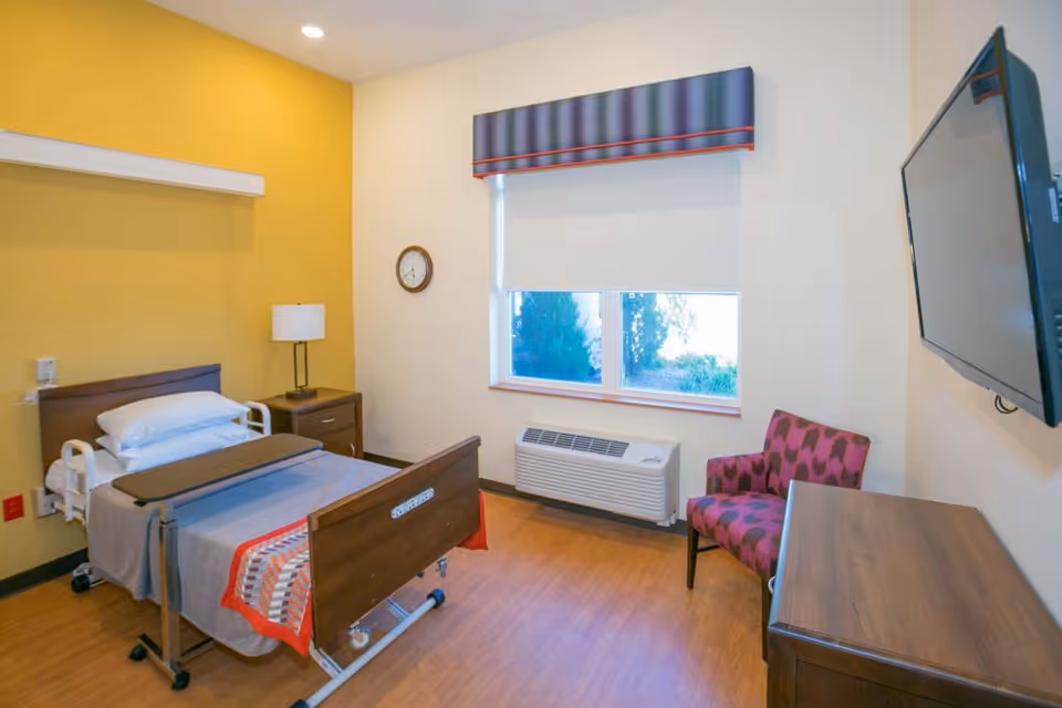 A clean and simple patient room with a hospital bed, bedside table with lamp, a wall clock, a window with a roller shade and valance, a patterned armchair, a wooden dresser, and a wall-mounted flat screen TV.