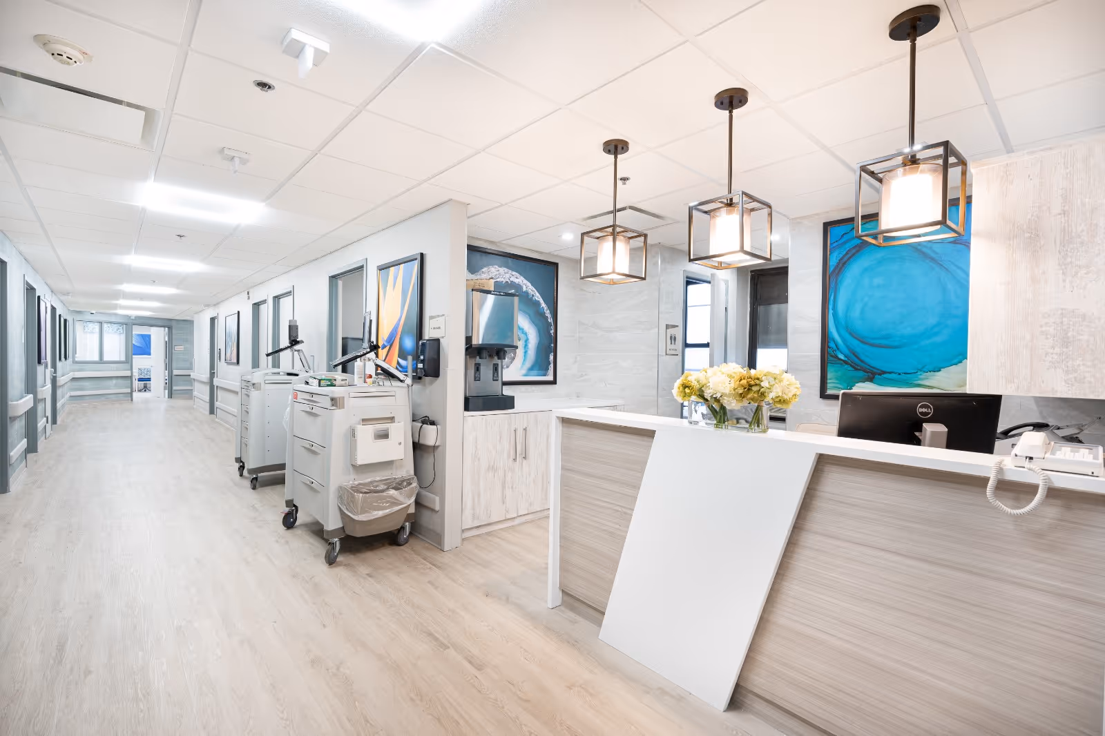 Reception desk and bright corridor inside a senior healthcare facility with medical carts, pendant lights, and blue wall art.