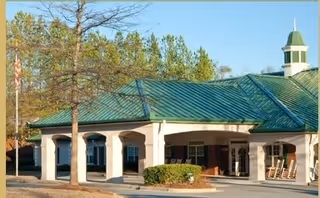 Exterior view of Ivy Hall Assisted Living facility showing a building with a green metal roof, white columns, and a small cupola on top. There are trees and shrubs around the building and an American flag on a flagpole to the left.