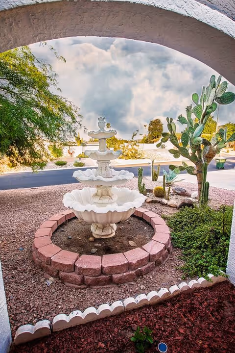 View of a white tiered water fountain surrounded by a circular brick border in a desert-style garden with gravel, cacti, and other desert plants, seen through a white stucco archway under a partly cloudy sky.