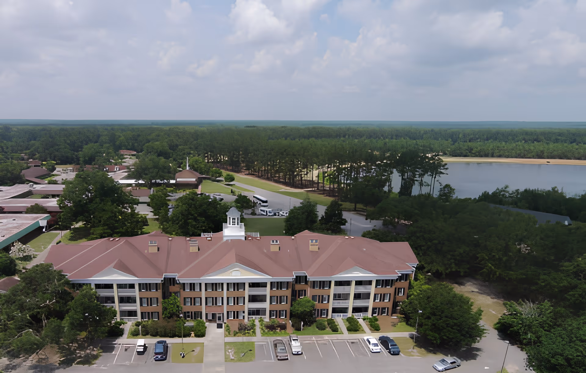 Aerial view of a large senior living facility building with a red roof, surrounded by trees and greenery. There is a parking lot with several cars in front of the building. In the background, there is a lake and a dense forest under a partly cloudy sky.