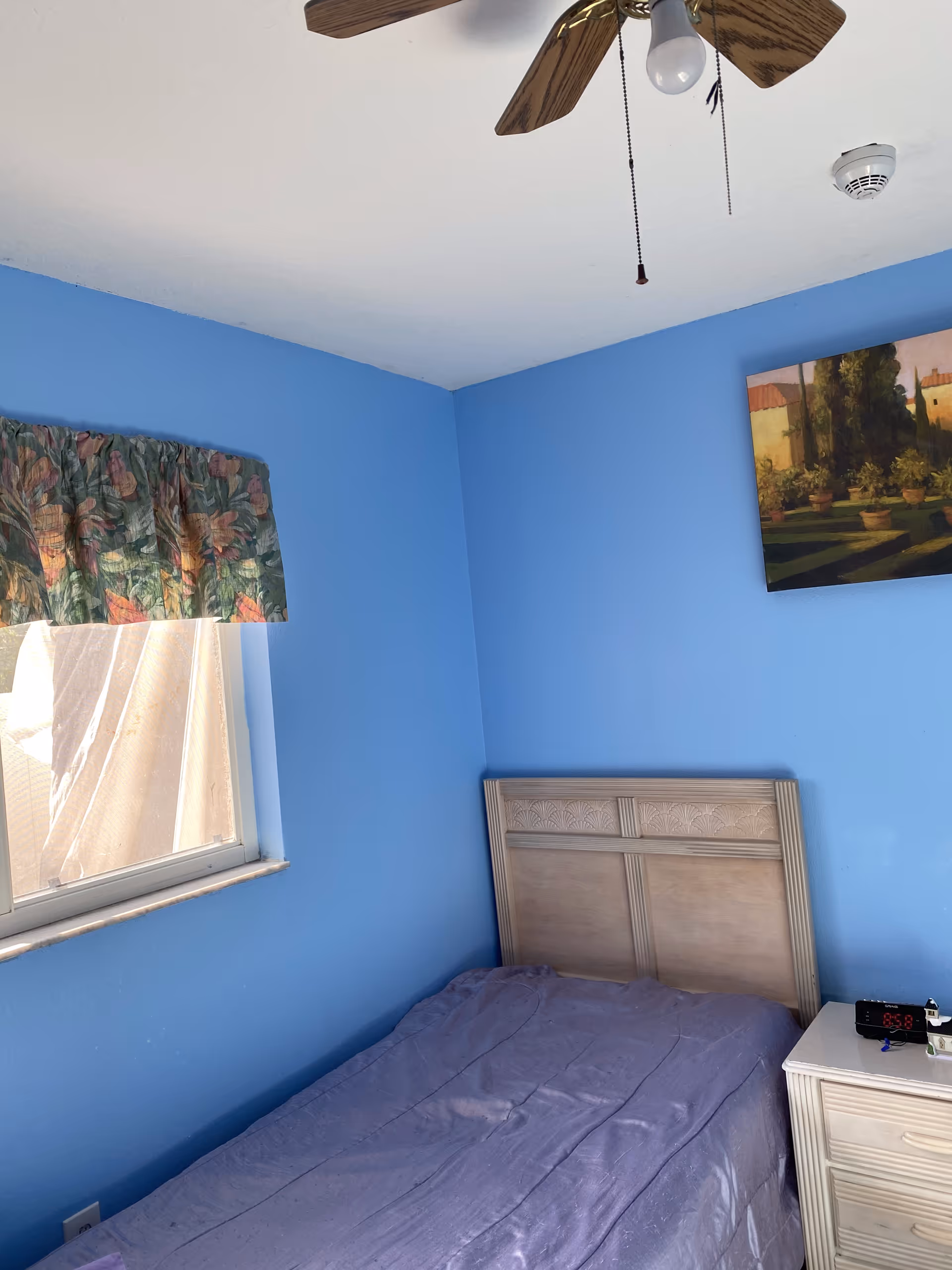 A small bedroom with blue walls, a window with floral curtains, a wooden headboard, a bed with a purple bedspread, a white nightstand with an alarm clock and small items, and a ceiling fan with wooden blades.