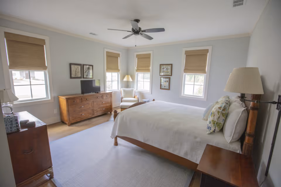 A bright and airy bedroom with a large bed covered in white bedding and floral pillows. The room features three windows with beige blinds, a wooden dresser with a small TV on top, a cozy armchair with a floor lamp beside it, and a ceiling fan overhead. The walls are painted light gray and the floor has a light-colored rug.