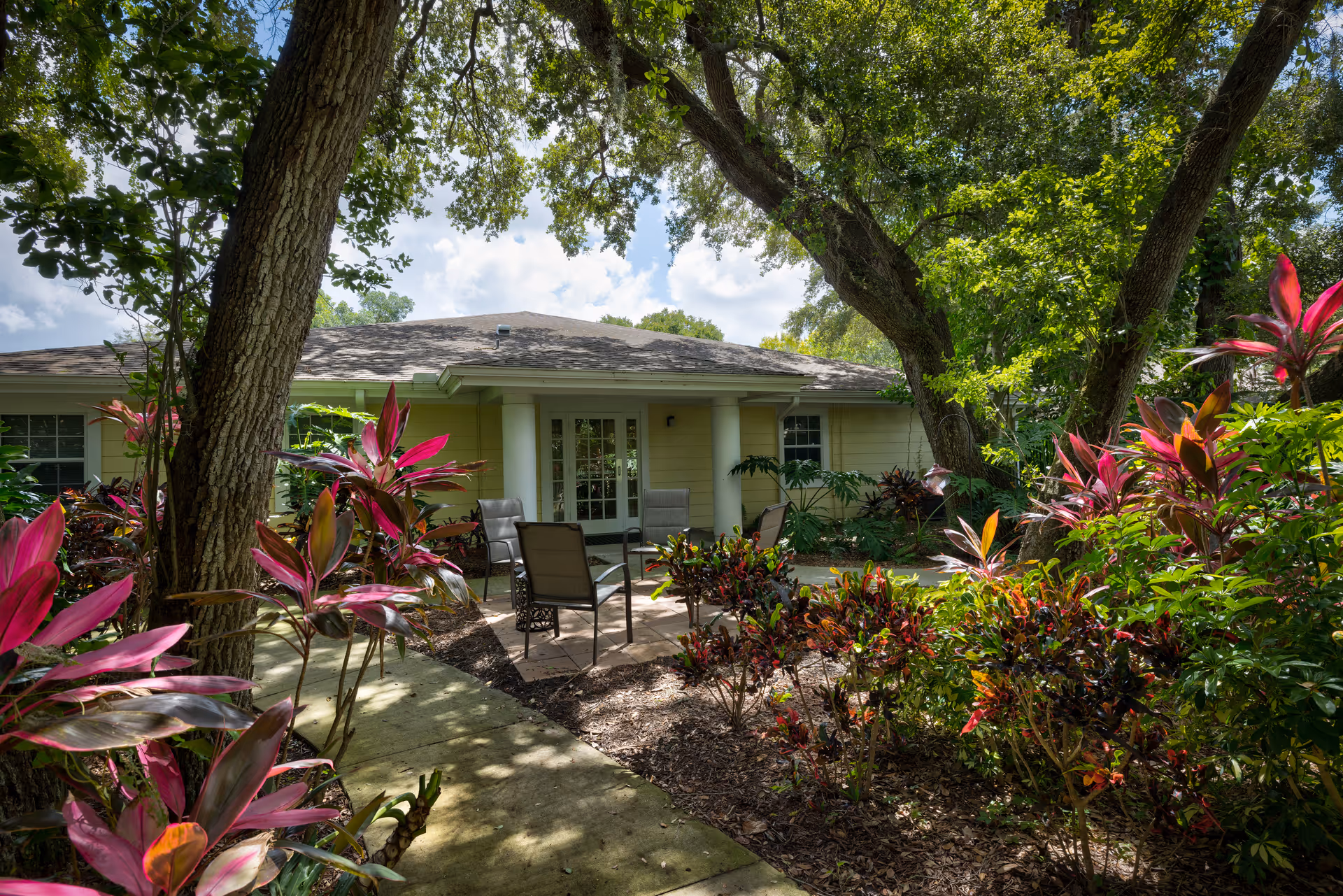 Outdoor patio area at Discovery Commons At Bradenton surrounded by lush greenery and colorful plants, with several chairs arranged around a small table under large trees providing shade.