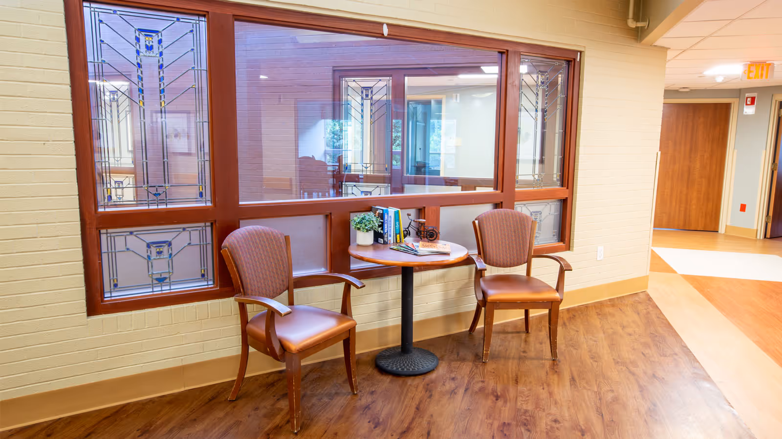 Two upholstered chairs and a small round table with books sit by decorative windows in a senior living facility hallway.