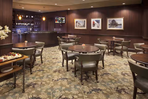 Cozy wood-paneled dining/lounge area with round tables, chairs, framed artwork, and a bar in the back.
