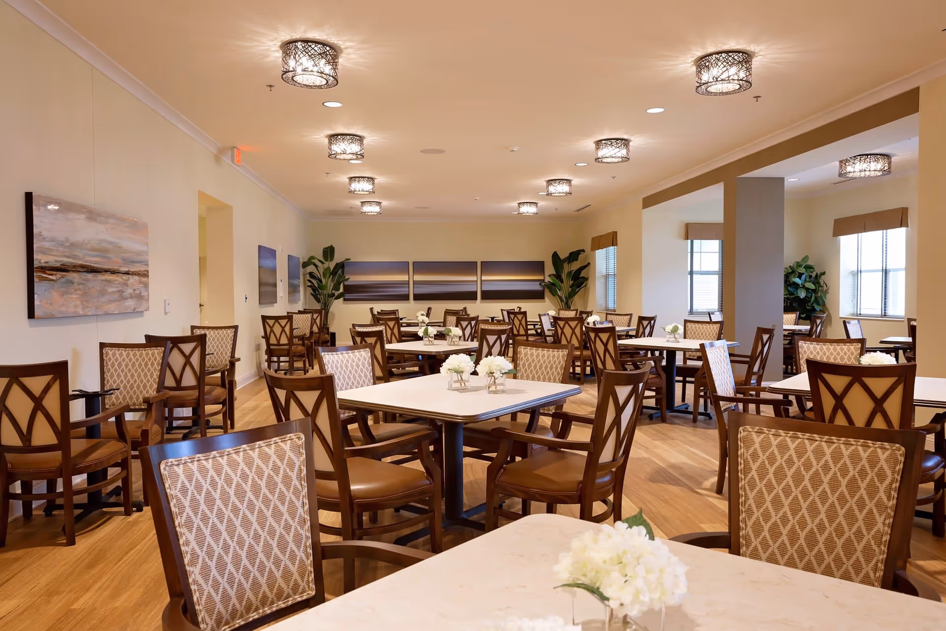 A spacious dining room with multiple square tables and wooden chairs with patterned upholstery. Each table has a small white floral centerpiece. The room has warm lighting from decorative ceiling fixtures, light-colored walls adorned with landscape paintings, and large windows with beige valances letting in natural light. There are also green potted plants in the corners.