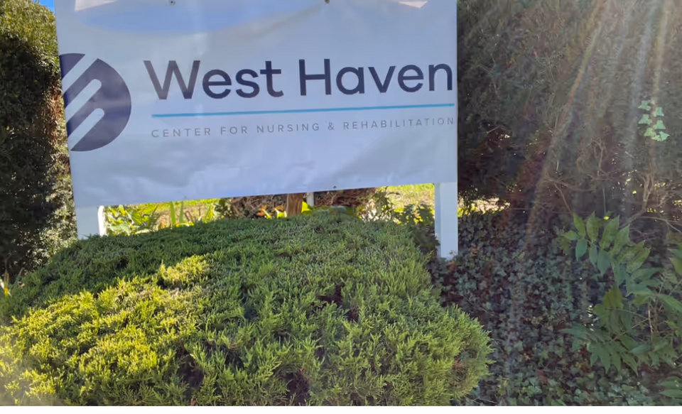 A yard sign reading "West Haven Center for Nursing & Rehabilitation" above trimmed shrubs with sunlight filtering through nearby bushes.