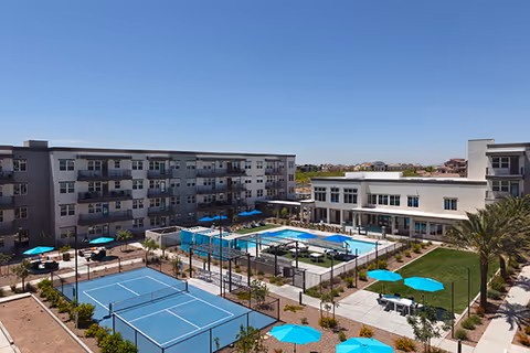 Outdoor view of a senior living facility named Mera Westgate featuring a swimming pool surrounded by lounge chairs and umbrellas, a tennis court, landscaped gardens, and multi-story residential buildings under a clear blue sky.