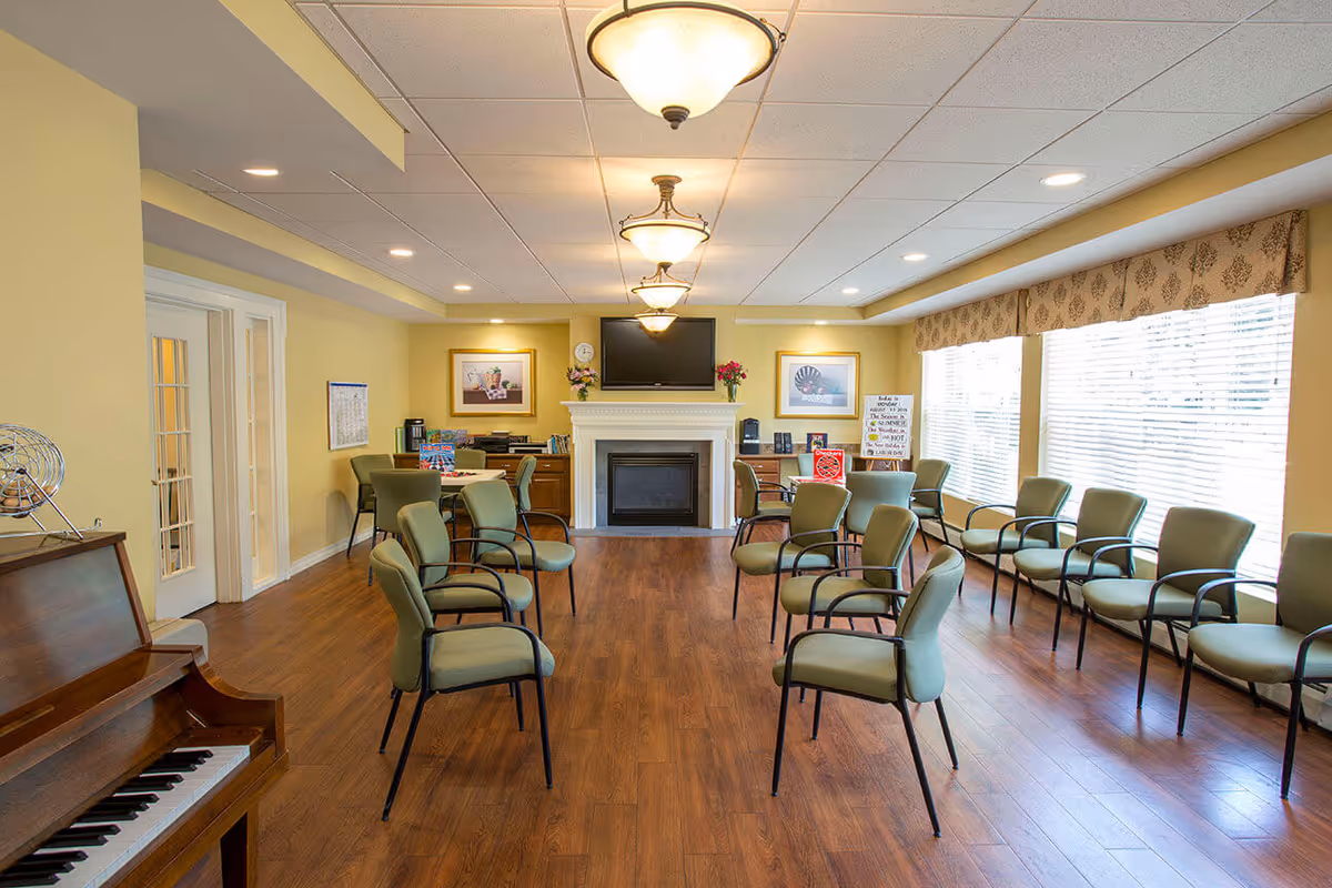 A spacious common room with wooden flooring and yellow walls, featuring multiple green cushioned chairs arranged in a semi-circle facing a white fireplace with a flat-screen TV mounted above it. The room is well-lit with ceiling lights and large windows covered with blinds and patterned valances. A piano is visible on the left side of the image, and there are framed pictures and flowers on the mantel and side tables.