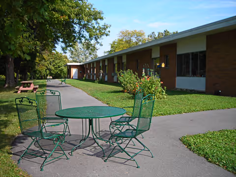 Outdoor area of Auburn Rehabilitation and Nursing Center featuring a paved walkway, green metal table with four matching chairs, picnic table, grass lawn, trees, and a single-story brick building in the background under a clear blue sky.
