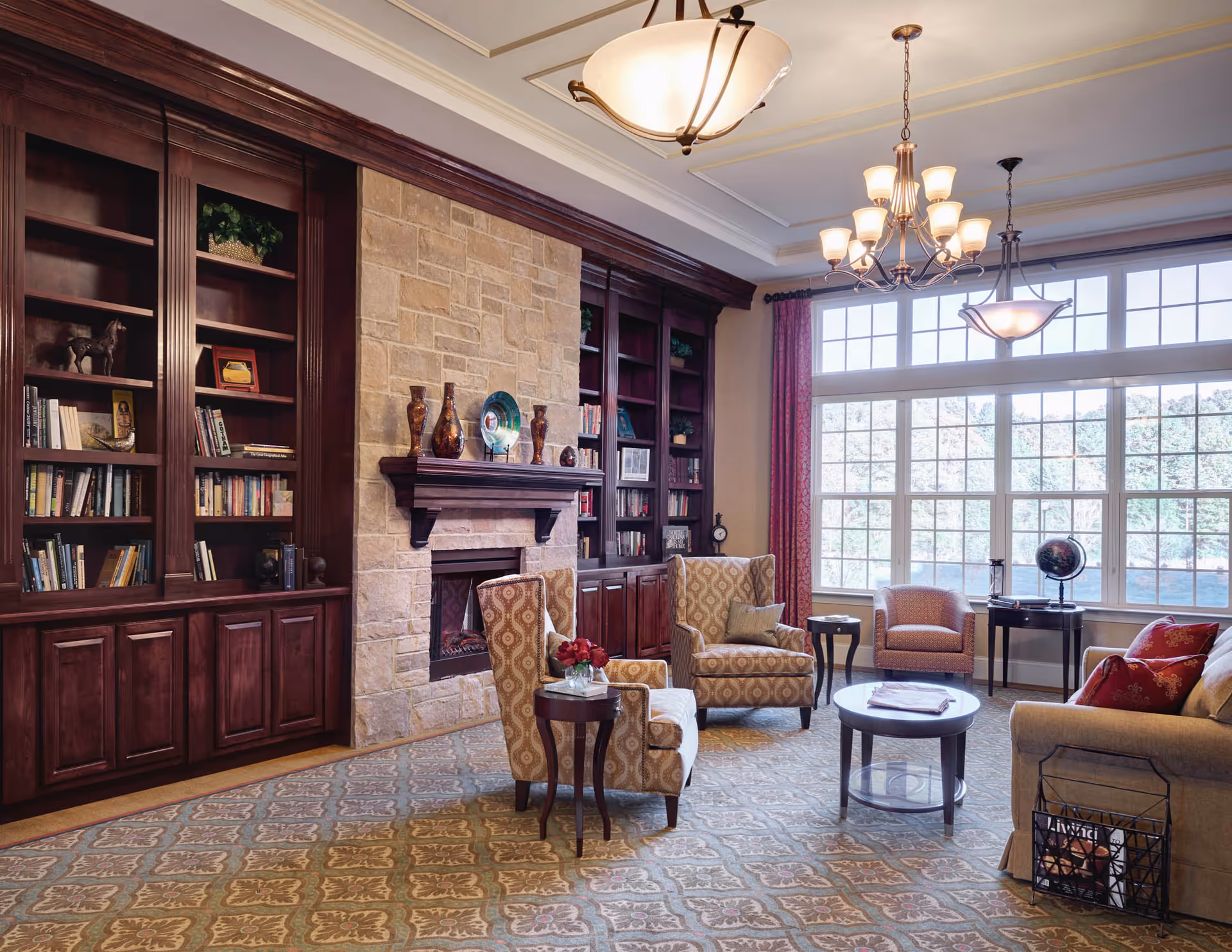Bright, cozy community living room with built-in bookshelves and a stone fireplace, upholstered armchairs, coffee tables, and large windows.