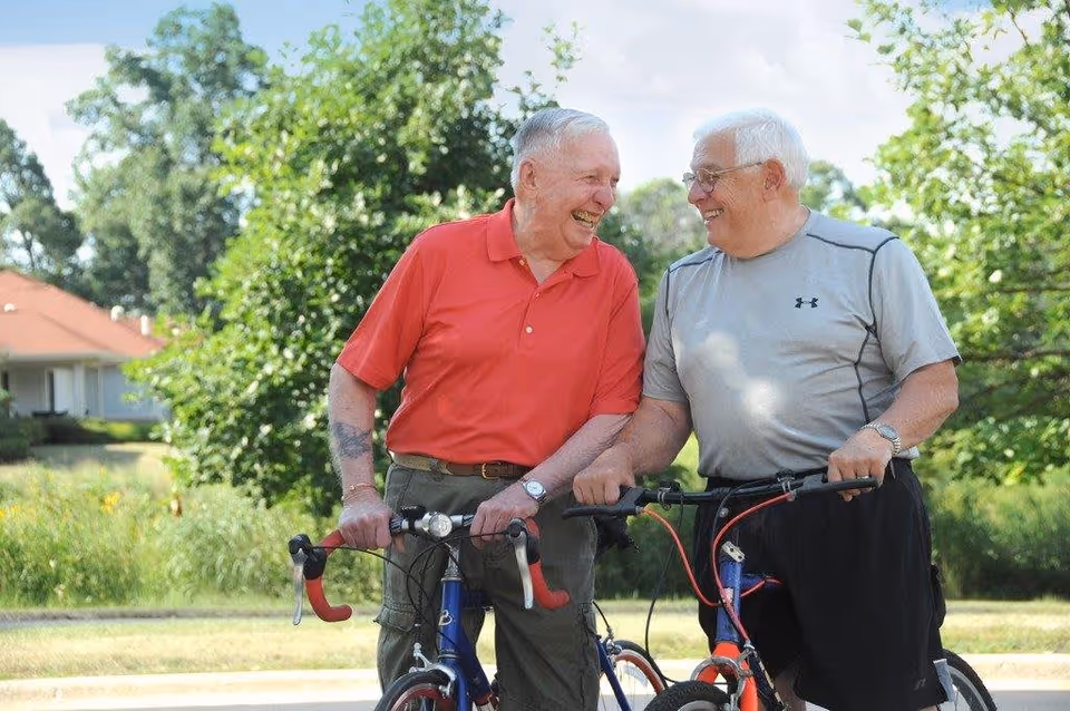 Two elderly men smiling and standing next to their bicycles outdoors on a sunny day, with trees and a house in the background.
