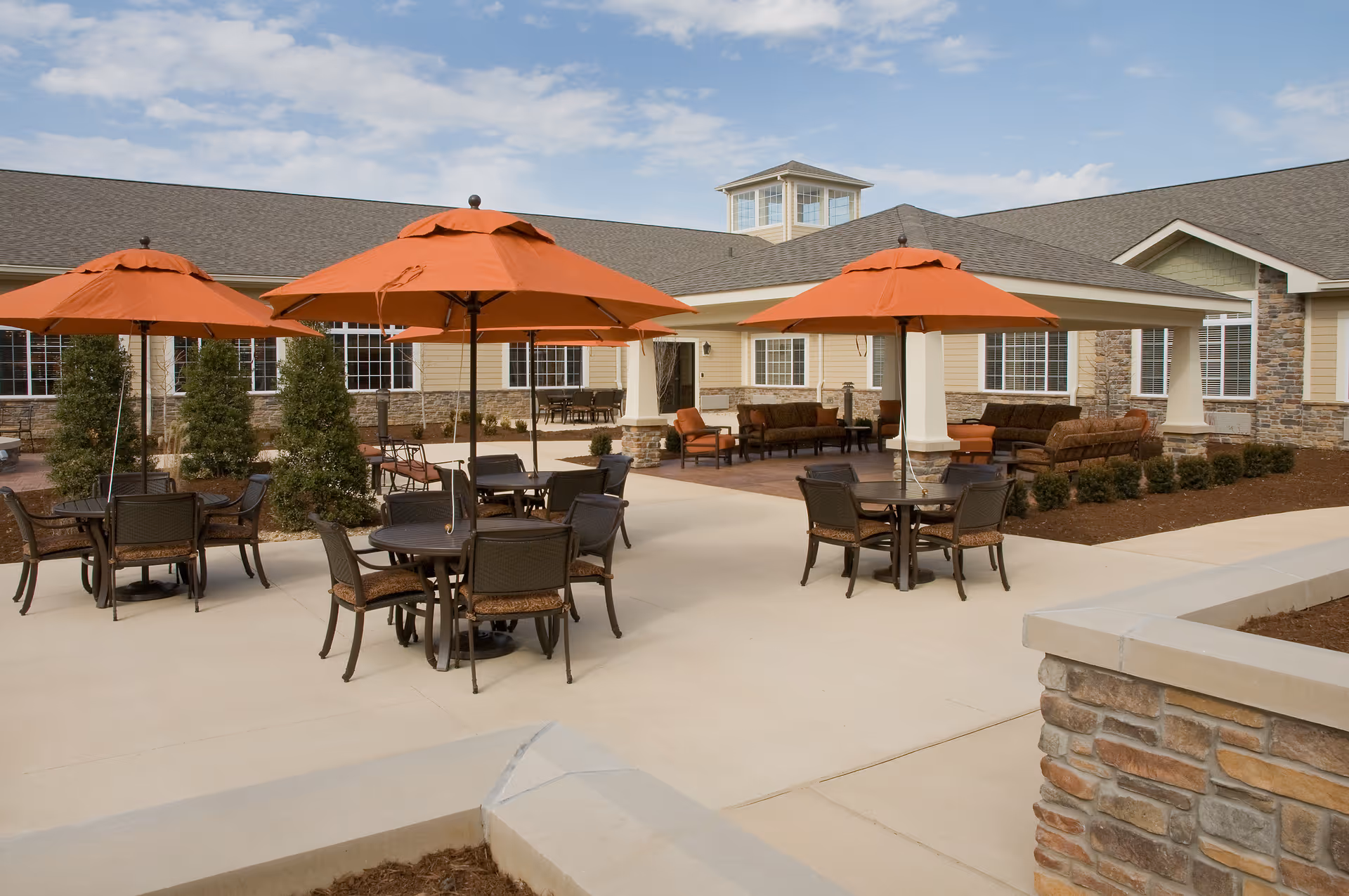 Outdoor patio area at a senior living facility with multiple round tables and chairs under orange umbrellas. The patio is surrounded by a building with beige siding and stone accents, featuring large windows and a covered seating area with cushioned chairs and sofas.