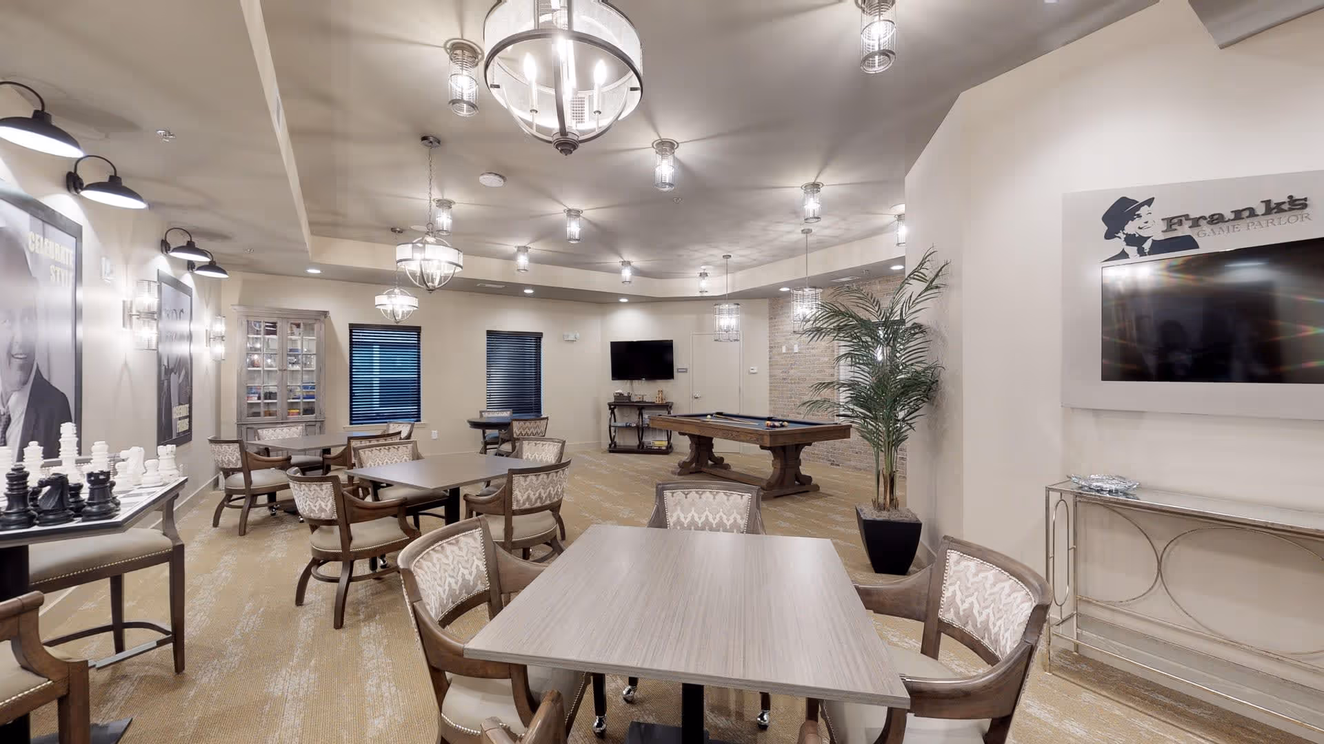A spacious game and lounge room with several tables and chairs arranged for group activities. There is a large chess set on a table to the left, a pool table in the back center, and a flat-screen TV mounted on the wall to the right with a sign above it reading 'Frank's Game Parlor.' The room is well-lit with multiple ceiling lights and decorated with framed pictures, a potted plant, and neutral-colored walls and carpet.