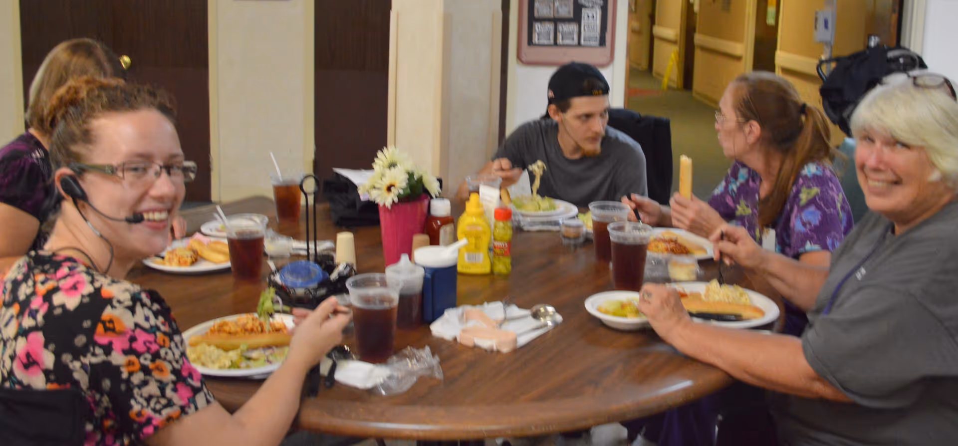 A group of five people sitting around a round wooden table enjoying a meal together. The table has plates of food, cups of iced tea, condiments, and a small flower arrangement in the center. The setting appears to be indoors with a hallway visible in the background.
