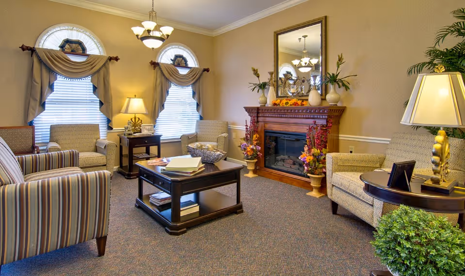 A cozy senior living facility lounge with multiple upholstered armchairs arranged around a dark wooden coffee table. The room features two arched windows with draped curtains, a wooden fireplace decorated with vases and flowers, a large mirror above the fireplace, and several lamps providing warm lighting. There is also a small side table with a lamp and a potted plant in the corner.