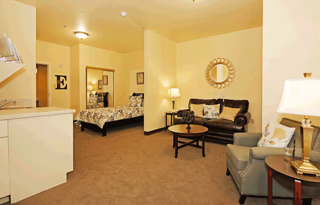 A cozy senior living facility room featuring a sleeping area with a bed covered in floral bedding, a sitting area with a dark leather loveseat, a gray armchair, a round wooden coffee table, and two side tables with lamps. A decorative round mirror hangs on the wall above the loveseat.