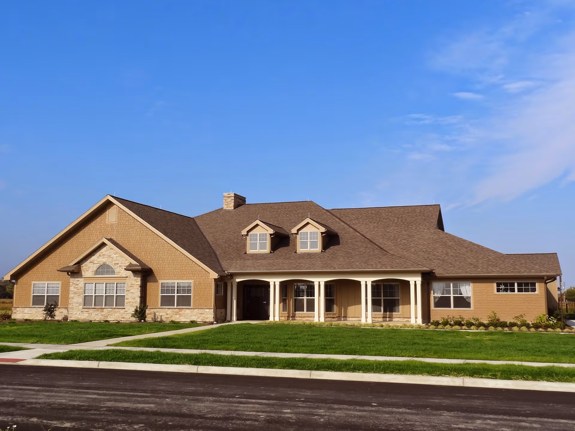 Exterior view of a single-story residential-style building with a brown roof and beige walls, featuring a front porch with columns, multiple windows, and a well-maintained lawn under a clear blue sky.
