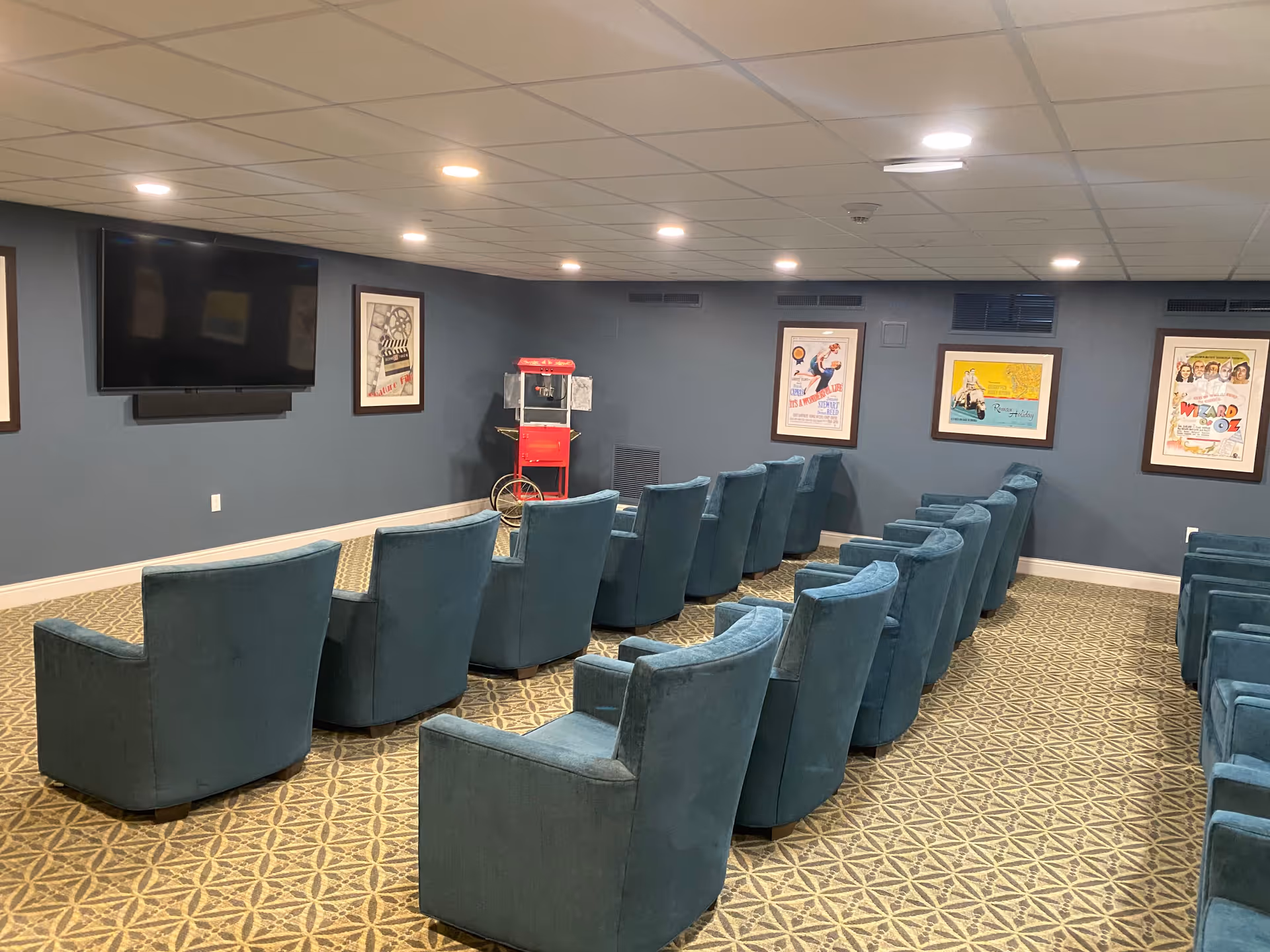 A small movie theater room with two rows of blue upholstered armchairs facing a large flat-screen TV mounted on a dark blue wall. The room has a patterned carpet, framed vintage movie posters on the walls, and a red popcorn machine in the corner.