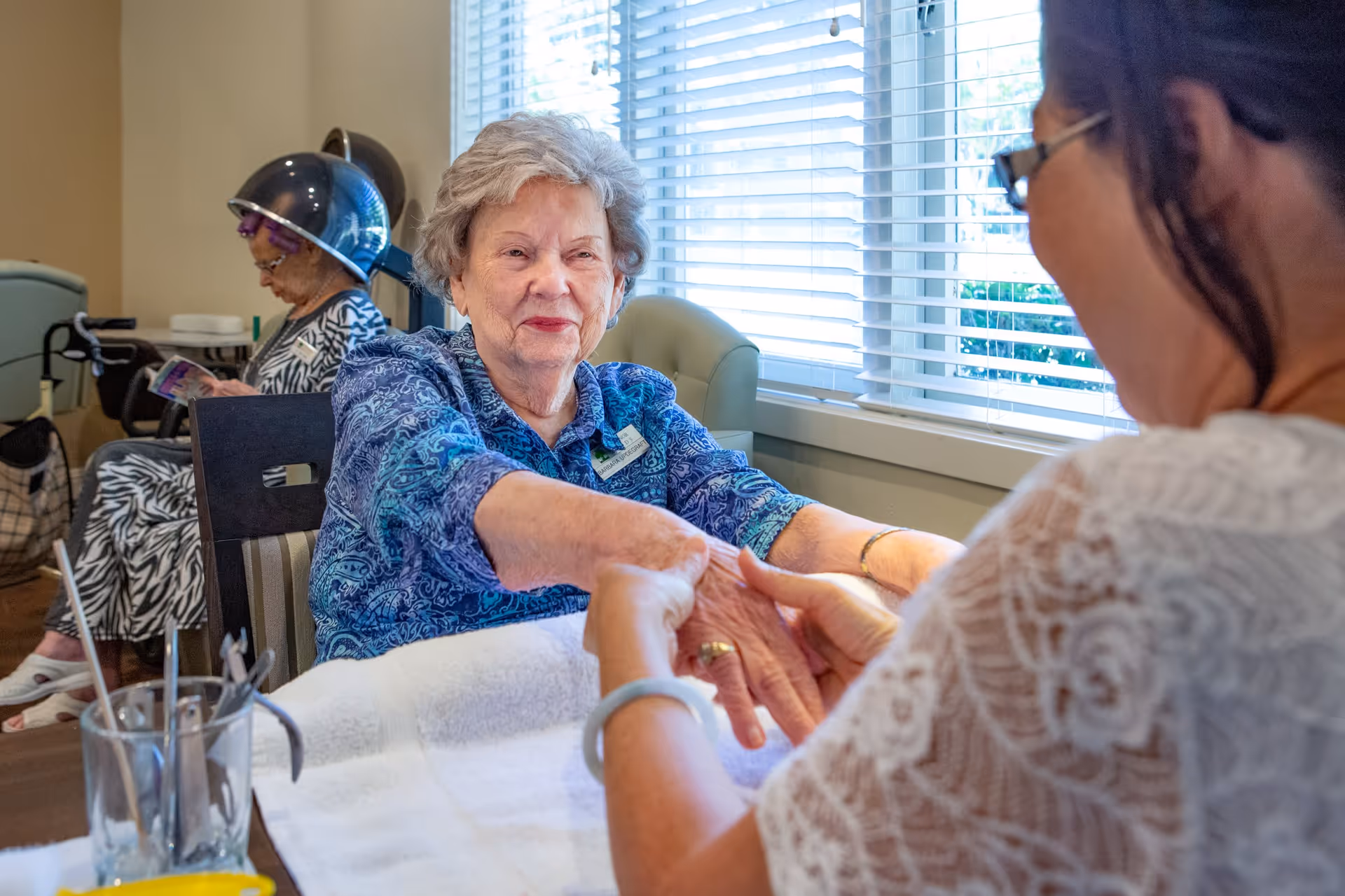 An elderly woman with gray hair wearing a blue patterned shirt is receiving a hand massage from another woman in a lace top. In the background, another elderly woman is sitting under a hair dryer, reading a magazine. The setting appears to be a bright room with large windows and blinds.