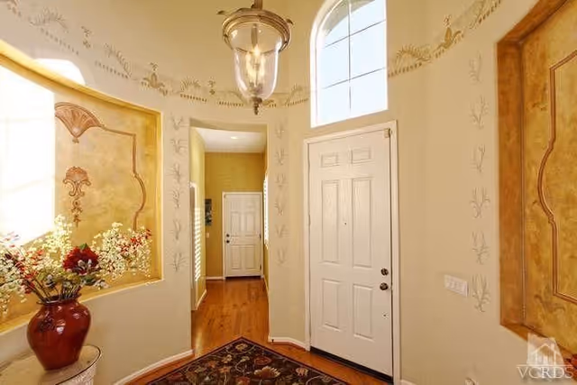 Bright foyer with a white front door, arched transom window, decorative wall niches, hardwood floor, and a vase of flowers.