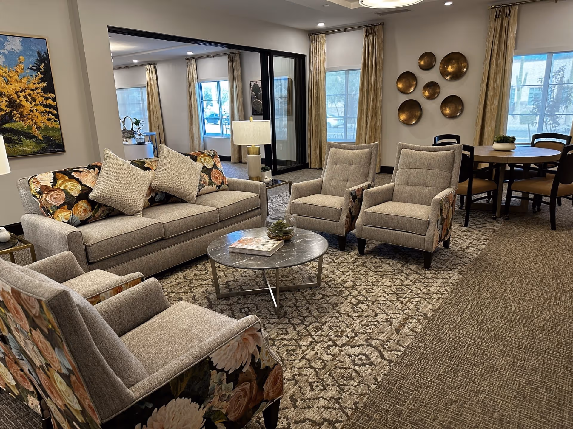 A cozy living room area in a senior living facility featuring a beige sofa with floral cushions, two matching armchairs with floral sides, a round marble coffee table with a book and a small plant, a round dining table with chairs in the background, large windows with beige curtains, and decorative wall art including a painting and metallic circular plates.