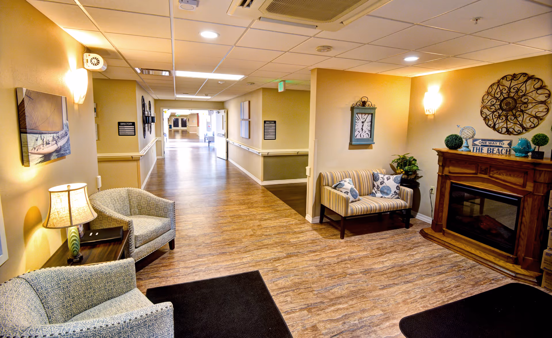 Well-lit senior living facility lobby with seating, a decorative fireplace, and a long hallway leading to other rooms.