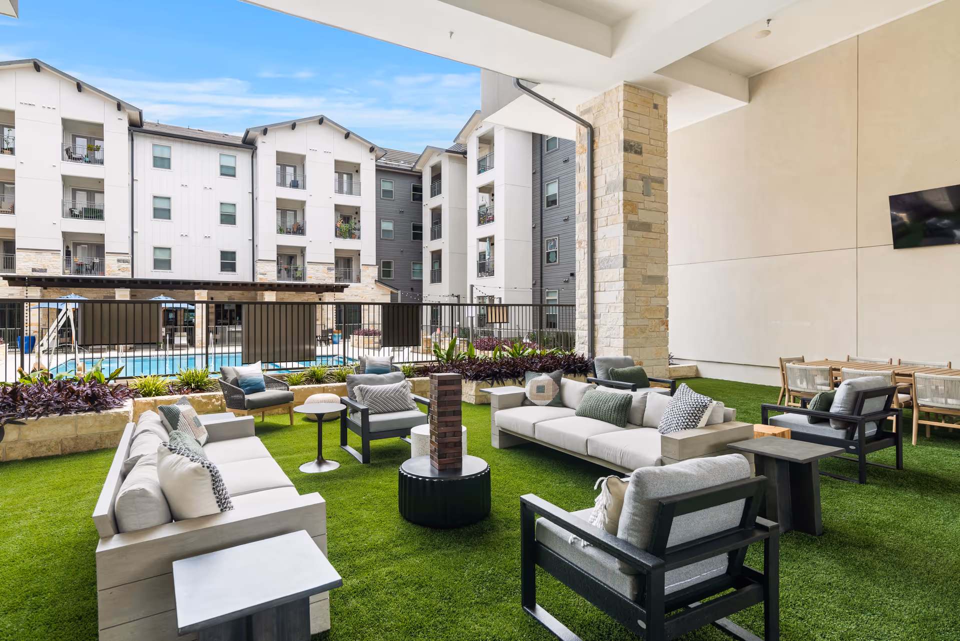Outdoor covered seating area with modern cushioned sofas and chairs arranged around a small table on artificial grass, overlooking a fenced swimming pool and multi-story residential building under a blue sky.