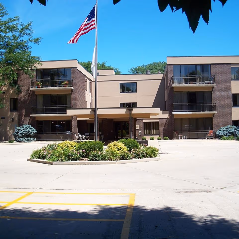 Front exterior view of a three-story senior living facility building with balconies, an American flag on a flagpole, and a landscaped circular garden in front of the entrance under a clear blue sky.