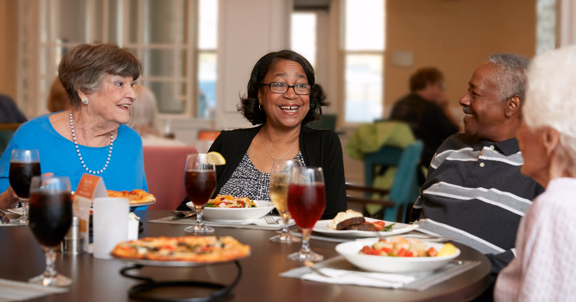 Four elderly people sitting around a dining table in a well-lit room, enjoying a meal together and engaging in conversation. The table has plates of food including pizza and salads, along with glasses of beverages.