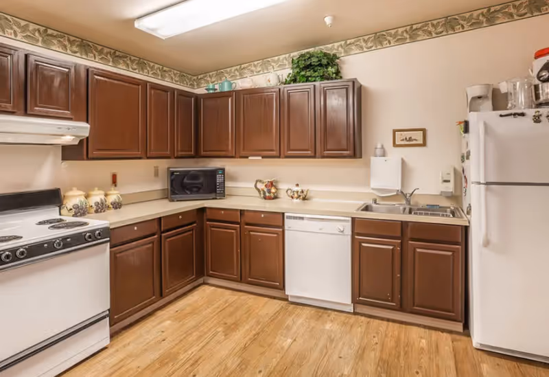 A kitchen with wooden cabinets, a white stove, a microwave, a dishwasher, a double sink, and a white refrigerator. The floor is wooden, and there is a floral wallpaper border near the ceiling.