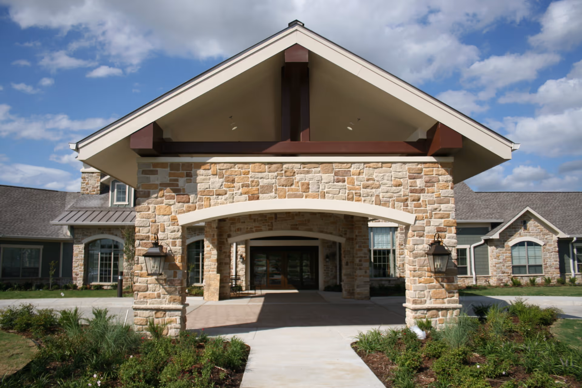Front exterior view of a building with stone facade and a large covered entrance supported by stone pillars, surrounded by landscaped greenery under a partly cloudy sky.