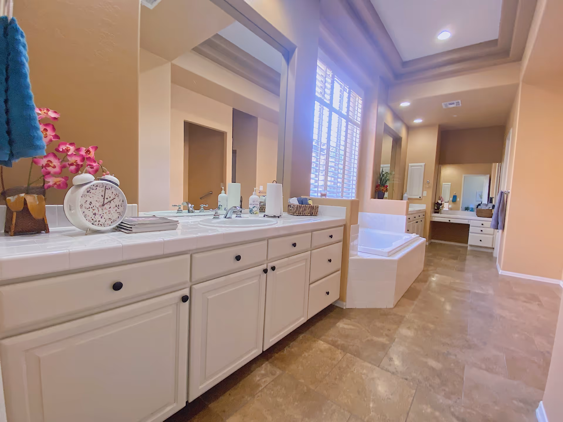 A spacious bathroom with beige walls and tiled floor featuring a long white vanity with two sinks, a large mirror, and various toiletries on the countertop. There is a built-in bathtub next to a window with white shutters, and recessed ceiling lights illuminate the room.