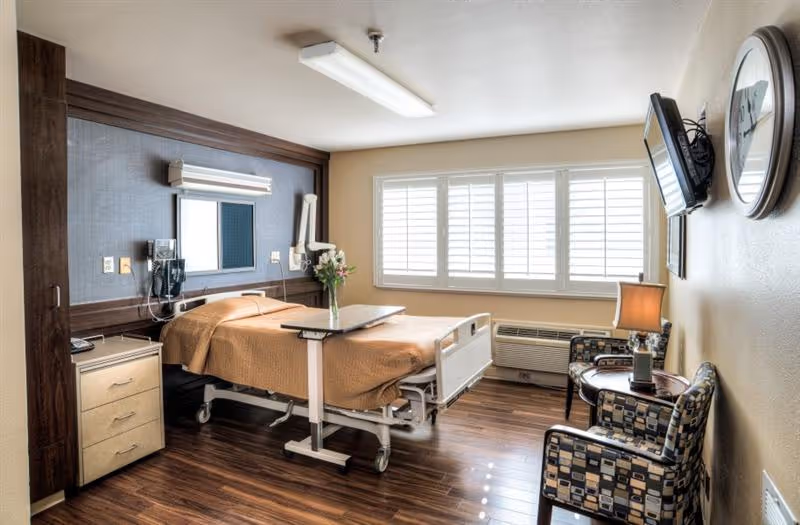 A well-lit healthcare facility room with a hospital bed covered in a brown blanket, a bedside table with drawers, a wall-mounted telephone, a large window with white shutters, two patterned armchairs, a small round table with a lamp, and a wall-mounted TV.