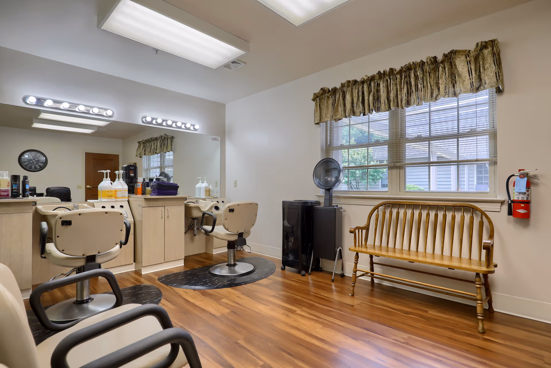 Interior of a senior living facility salon with two beige salon chairs in front of a large mirror and countertop with hair care products. There is a wooden bench under a window with curtains, a hair dryer, and a fire extinguisher mounted on the wall. The floor is wooden and the room is well-lit with ceiling lights.